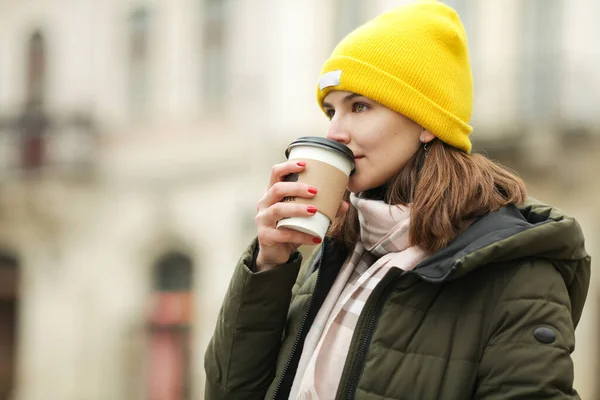 コーヒー愛好家のコンセプト ヨーロッパの街で熱い飲み物を飲む若い女性 緑のコート 白とベージュのスカーフ 黄色のビーニーを身に着けているモデル 閉めろ 屋外撮影 — ストック写真