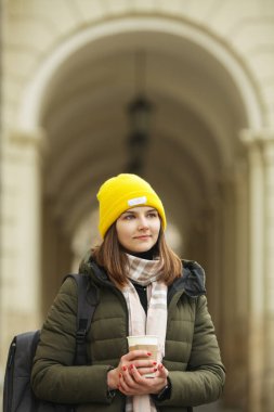 Seyahat ve kahve konsepti: Avrupa 'nın caddesinde sıcak içecek içen güzel gülümseyen genç bir kadının portresi. Yeşil ceketli, beyaz ve bej atkılı bir manken. Açık hava çekimi