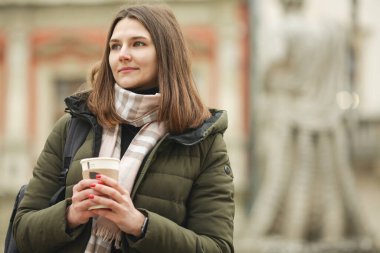 Kahve aşığı kavramı: gülümseyen genç kadın Avrupa 'nın caddesinde sıcak içecek içiyor. Yeşil ceketli, beyaz ve bej atkılı bir manken. Kapatın. Kopyalama uzayı. Açık hava çekimi