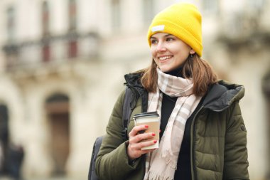 Coffee lover concept: happy smiling young woman drinking hot beverage at street of European city. Model wearing green coat, white, beige scarf, yellow beanie. Close up. Text space. Outdoor shot