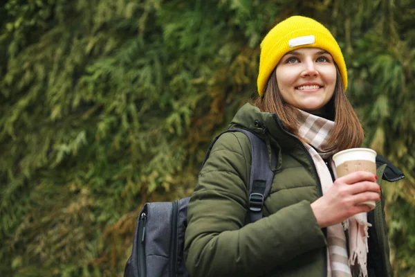 咖啡爱好者的概念 快乐微笑的年轻女子喝着热饮料参观国家公园 穿着绿色外套 米黄色围巾 黄色便帽的模特 靠近点文字空间 室外开枪 — 图库照片