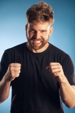 Spor zaferi konsepti. Çığlık atan karizmatik, aktif, 30 yaşında, mavi arka planda elleri havada poz veren birinin duygusal portresi. Mükemmel saç kesimi. Hippi tarzı. Beyaz, parlak bir gülümseme. Stüdyo çekimi