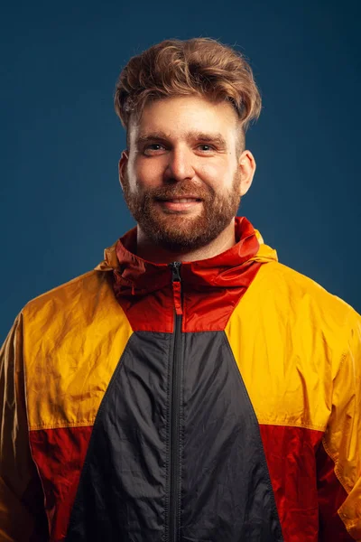 観光やスポーツのコンセプトのためのアウター 青色の背景に立ってカリスマ筋肉30歳の男性笑顔の感情的な肖像画 スタイリッシュなヘアカット カラフルなジャケット スタジオショット — ストック写真
