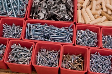 Tahta bir arka planda kırmızı plastik tepsi kutusunda çeşitli boyutlarda vida, cıvata, tahta çiviler ve tahta çiviler var. Mobilyalar için montaj seti. Seçici odak.