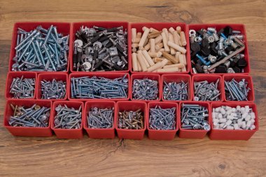 Tahta bir arka planda kırmızı plastik tepsi kutusunda çeşitli boyutlarda vida, cıvata, tahta çiviler ve tahta çiviler var. Mobilyalar için montaj kiti.