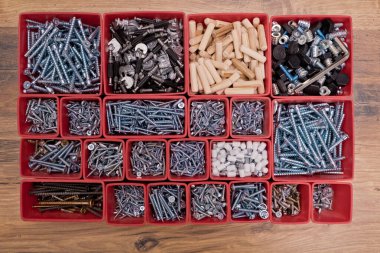 Tahta bir arka planda kırmızı plastik tepsi kutusunda çeşitli boyutlarda vida, cıvata, tahta çiviler ve tahta çiviler var. Mobilyalar için montaj seti. Üst görünüm.