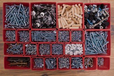 Tahta bir arka planda kırmızı plastik tepsi kutusunda çeşitli boyutlarda vida, cıvata, tahta çiviler ve tahta çiviler var. Mobilyalar için montaj seti. Üst görünüm.