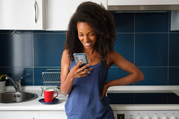 Casual afro hairstyle woman at home using smartphone for video call from the kitchen. Cheerful female searching cocking recipes online concept.