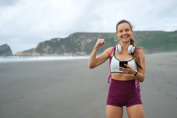 健身锻炼的动机和成功 身材魁梧的女人在海滩用手机应用程序庆祝锻炼的成就 — 图库照片