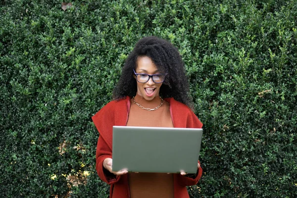令人惊讶的时髦黑人妇女使用笔记本电脑 秋季时尚网上购物理念 在外面拿着电脑的有表情的女人 图库照片