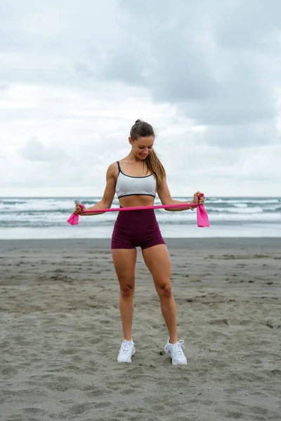 适合运动的年轻女子在海滩上锻炼 女运动员使用弹性带训练腿的力量 户外运动健身 图库图片