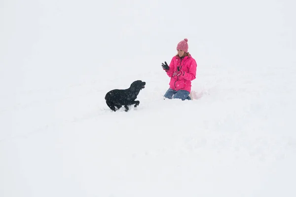 穿着粉色夹克的年轻女人在外面雪地里和她的黑色拉布拉多猎犬玩耍 — 图库照片