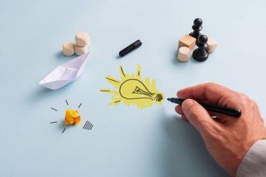 Satranç figürleri, origami teknesi ve zarların yanında mavi arka planda siyah kalemli bir ampul çizen bir iş adamının eli..