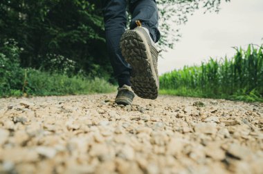 Low angle view of a male legs in hiking boots walking or running on a gravel country road with green corn fields around.