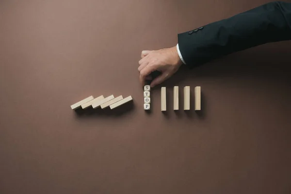 Hand of a businessman stacking wooden cubes with a Stop sign on them in the middle of collapsing dominos to prevent a business crisis.