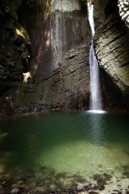 Kozjak waterfall in slovenia, europe in an open top cave with hight rocky walls and green lake.