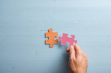 Male hand matching two blank puzzle pieces in orange and pink pastel colours. Over blue wooden background.