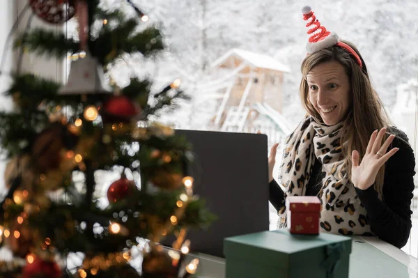 快乐的年轻女性头戴圣诞礼帽 兴奋地走向笔记本电脑 通过在线视频电话与家人或朋友见面 在家里旁边 有一棵装饰过的节日树 外面有冬天的自然气息 — 图库照片