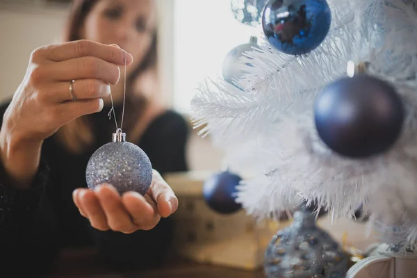 年轻女子手里拿着闪闪发亮的蓝色节日礼物 正要挂在一棵白色的圣诞树上 — 图库照片