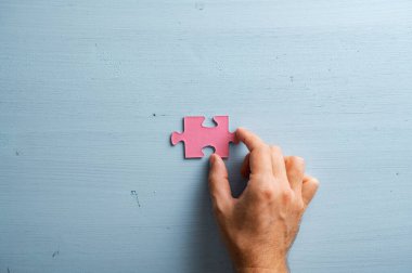 Erkek el, boş bir pembe yapboz parçasını sade mavi ahşap arka plana yerleştiriyor..