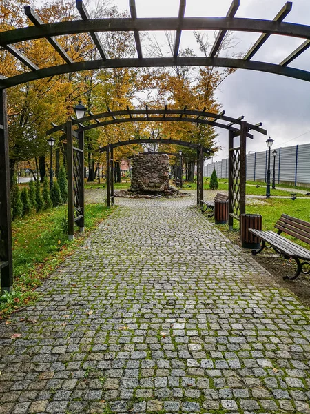 Small wooden pergola above small path in park with benches and bushes around