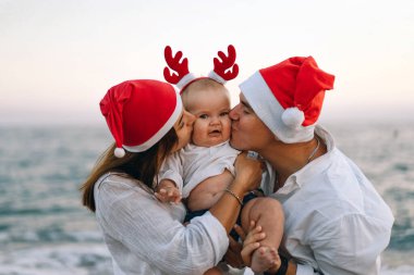 Kırmızı Noel Baba şapkalı bir aile sahilde yürüyor. Aile Noel 'i ve yeni yılı tropiklerde kutluyor..