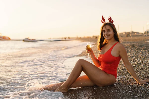 Noel Baba şapkalı genç ve güzel bir kadın elinde bir bardak şampanyayla sahilde yürüyor.. — Stok fotoğraf