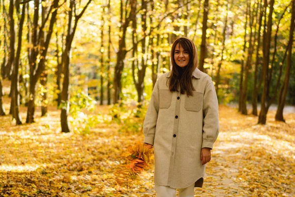 Woman in a long shirt with a bouquet of autumn leaves in her hands walks through the autumn forest.