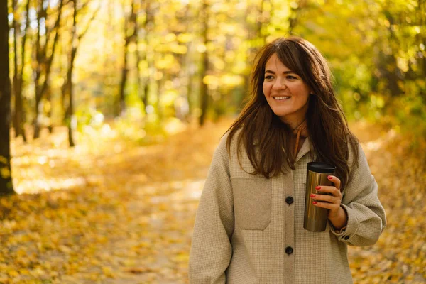 一个穿着长衫、手里拿着热杯子的年轻女子穿过秋天的森林. — 图库照片