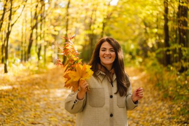 Ellerinde bir buket sonbahar yaprağı olan uzun tişörtlü bir kadın sonbahar ormanında yürüyor..
