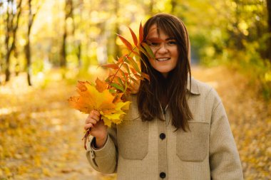 Ellerinde bir buket sonbahar yaprağı olan uzun tişörtlü bir kadın sonbahar ormanında yürüyor..