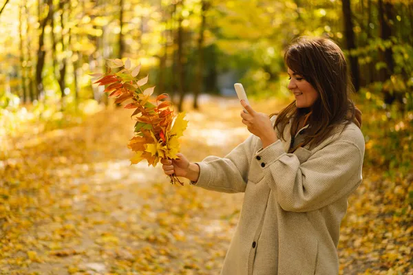 Frau im langen Hemd fotografiert Blätter am Telefon im Herbstwald. Naturlandschaft — Stockfoto