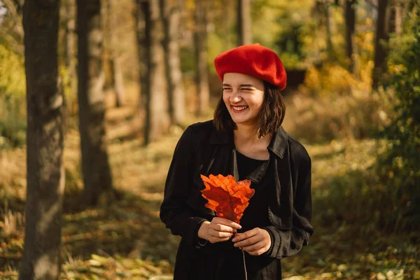 Теенгірл у червоному береті з осіннім листям у руках ходить лісом. — стокове фото