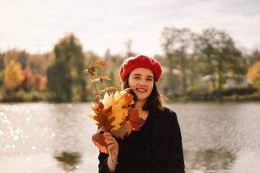 Elinde bir buket sonbahar yaprağı olan kırmızı bereli bir genç kız ormanda yürüyor.