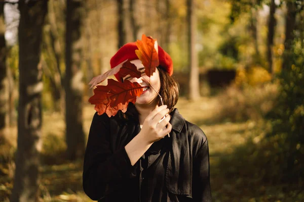 Teengirl punaisessa baskerissa syksyn lehtien kimppu käsissään kävelee metsän läpi. — kuvapankkivalokuva