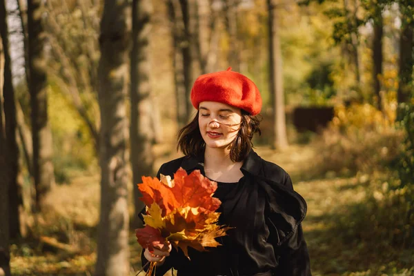 Una adolescente en una boina roja con un ramo de hojas de otoño en sus manos camina por el bosque — Foto de Stock
