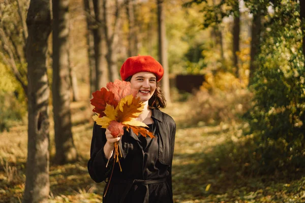 Ein Teengirl mit roter Baskenmütze und einem Strauß Herbstblätter in der Hand geht durch den Wald — Stockfoto