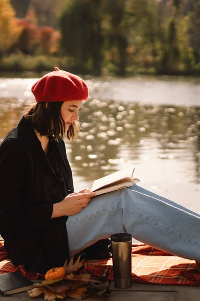 A Teengirl in a red boet reading book on wooden pontoon. Temporada de otoño. — Foto de Stock