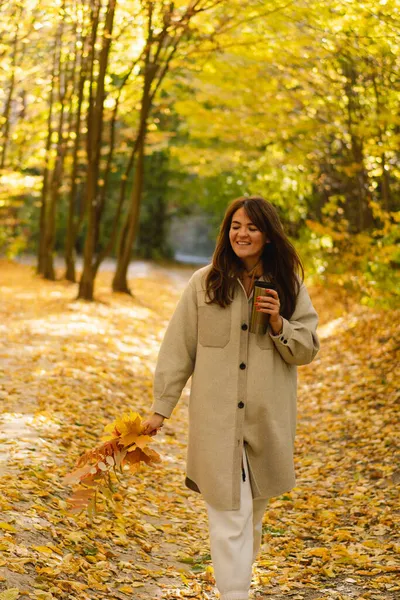 一个穿着长衫、手里拿着热杯子的年轻女子穿过秋天的森林. — 图库照片