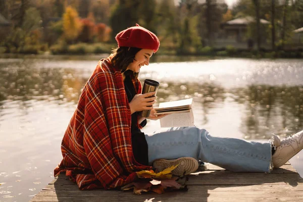 Uma adolescente em uma boina vermelha lendo livro sobre pontão de madeira. Temporada de outono. — Fotografia de Stock