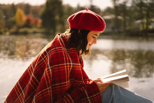 Uma adolescente em uma boina vermelha lendo livro sobre pontão de madeira. Temporada de outono. — Fotografia de Stock