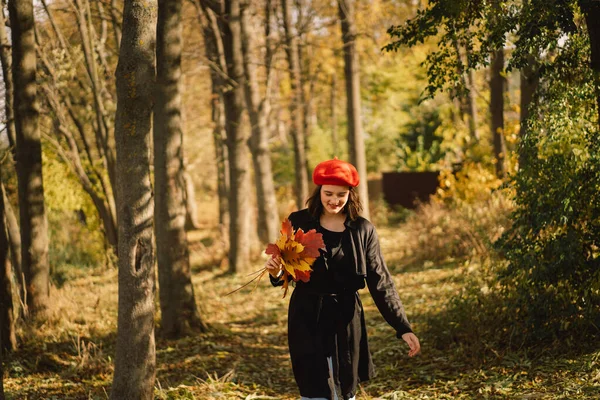 Ein Teengirl mit roter Baskenmütze und einem Strauß Herbstblätter in der Hand geht durch den Wald — Stockfoto