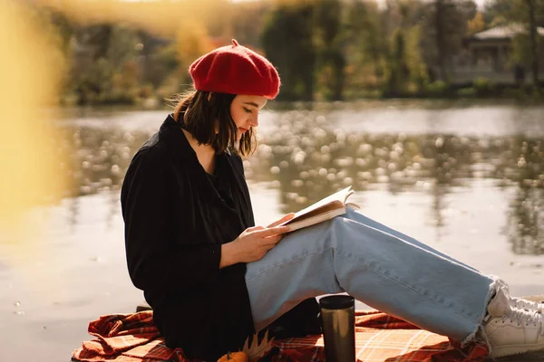 Sebuah Teengirl di baret merah membaca buku di ponton kayu. Musim gugur. — Stok Foto