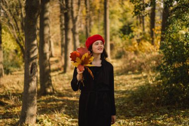 Elinde bir buket sonbahar yaprağı olan kırmızı bereli bir genç kız ormanda yürüyor.