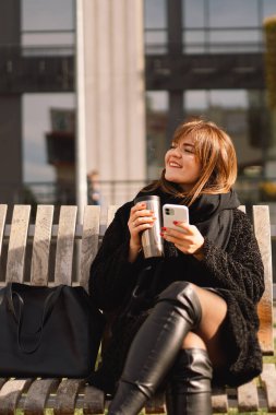 Şehirdeki bir bankta oturan bir kadın termo bardaktan çay içiyor ve telefon kullanıyor..