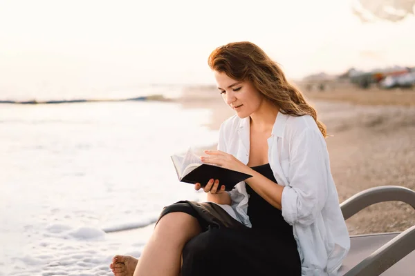 La mujer tiene el libro en sus manos. Leyendo el libro sobre un mar al atardecer. — Foto de Stock