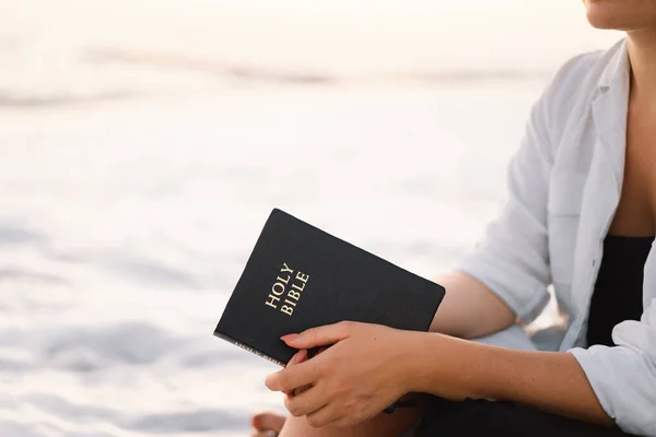La mujer cristiana tiene la Biblia en sus manos. Leyendo la Santa Biblia en el mar durante el hermoso atardecer — Foto de Stock