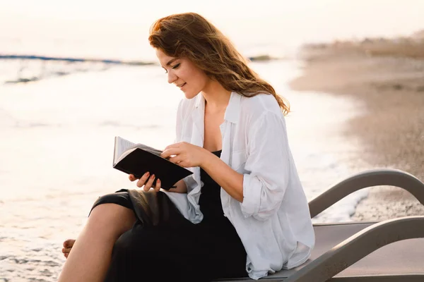 La mujer tiene el libro en sus manos. Leyendo el libro sobre un mar al atardecer. — Foto de Stock