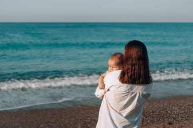 Bezli küçük bir çocukla kumsalda yürüyen genç bir anne..