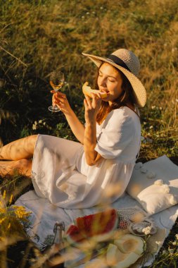 Beyaz elbiseli genç bir kız bir bardak şampanya ya da beyaz şarapla piknik yapıyor.